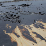 Derrame de petróleo en la costa, con manchas negras y aceitosas sobre la arena y el agua, símbolo de contaminación marina.