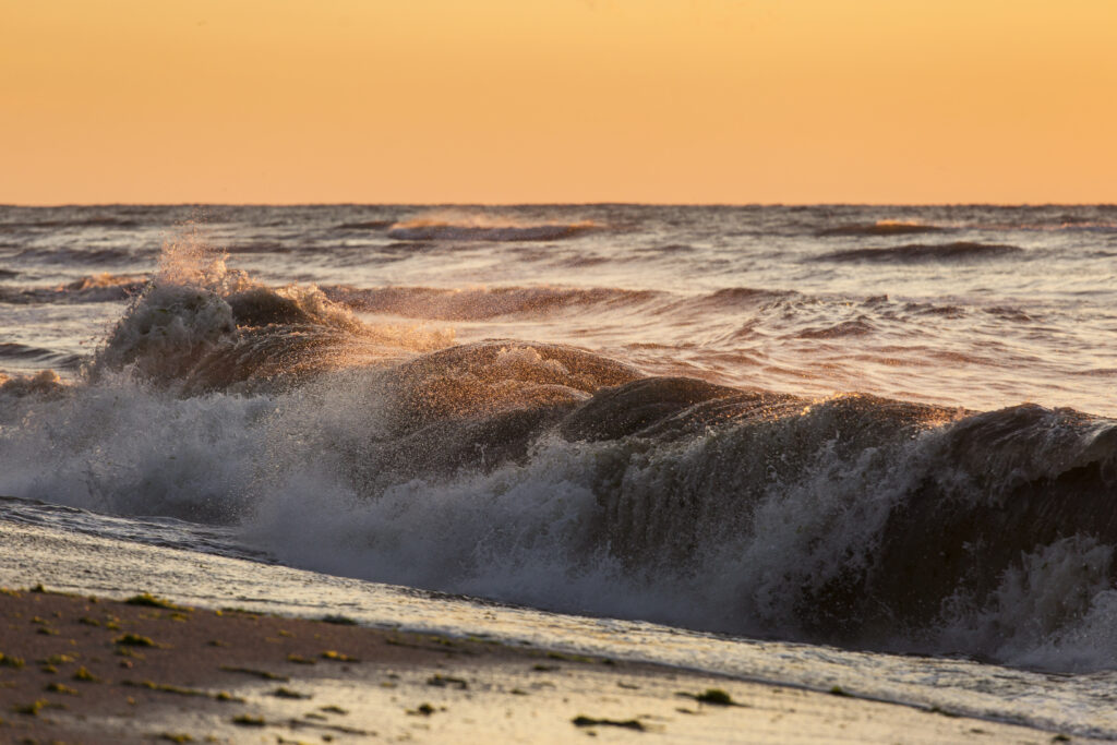 Olas agitadas rompiendo en la orilla durante un atardecer, reflejando tonos cálidos del sol sobre el agua.