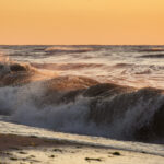 Olas agitadas rompiendo en la orilla durante un atardecer, reflejando tonos cálidos del sol sobre el agua.