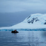 Grupo de científicos a bordo de una pequeña embarcación frente a un glaciar en la península Antártica, con montañas nevadas al fondo.