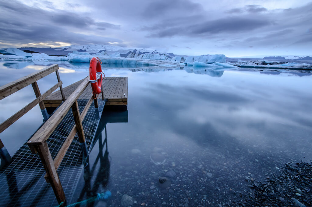 Un muelle de madera con salvavidas naranja sobresale sobre aguas calmas y gélidas, con témpanos de hielo flotando al fondo en un paisaje polar.