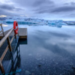 Un muelle de madera con salvavidas naranja sobresale sobre aguas calmas y gélidas, con témpanos de hielo flotando al fondo en un paisaje polar.