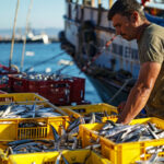 Hombre revisando cajas llenas de peces recién capturados sobre un muelle pesquero, con barcos al fondo y el mar de fondo.