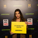 Mujer sostiene cartel amarillo con la frase "SALMONES POR AQUÍ NO" frente a un fondo con logos de Greenpeace y History Channel, en el marco de una campaña ambiental.